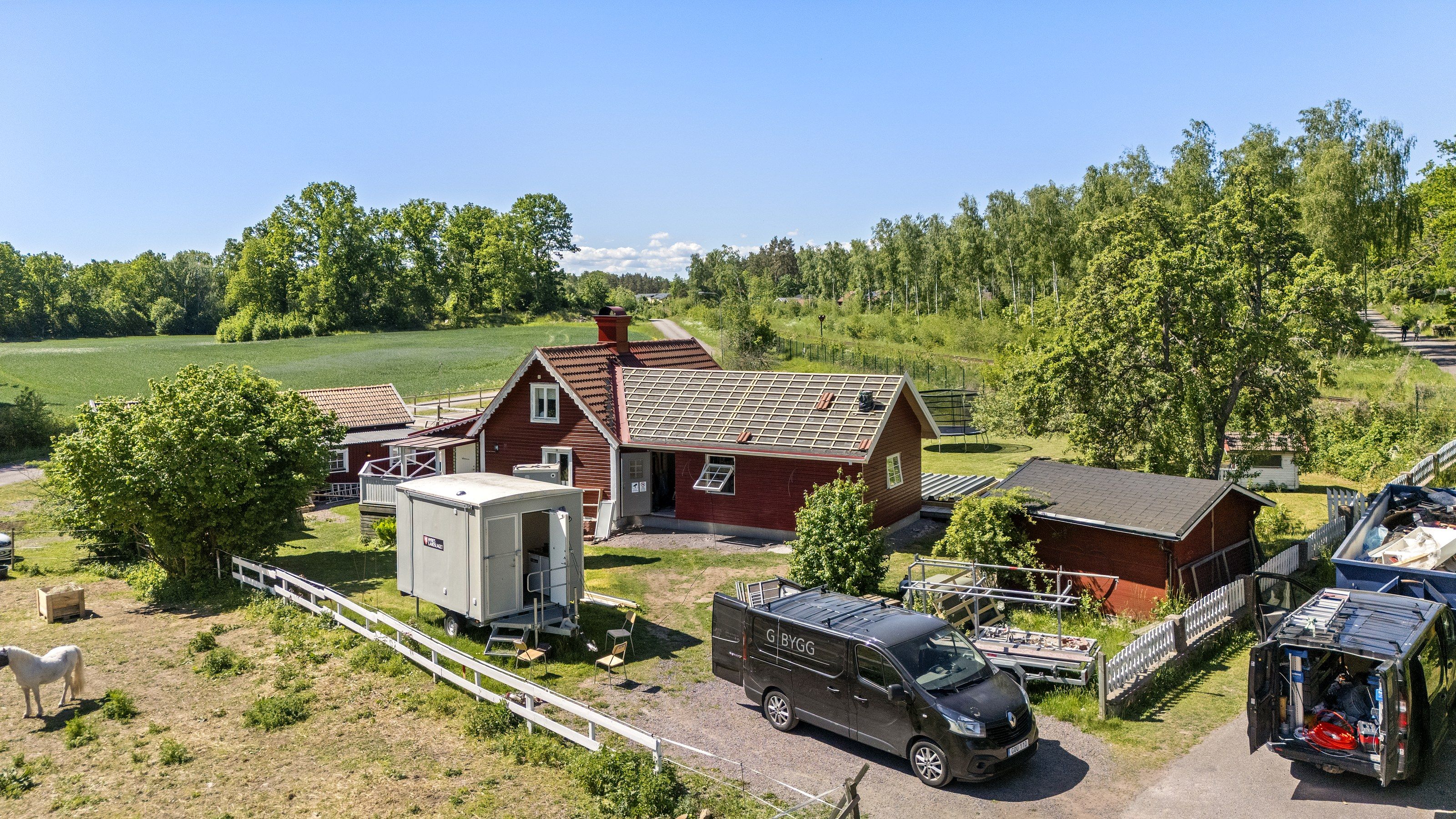 G-Byggs svarta servicebilar och en arbetsbod vid ett rött trähus vars tak läggs om, sett från ovan